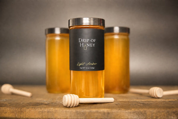 Three 12 oz jars of Drip of Honey on a wood table with honey dippers
